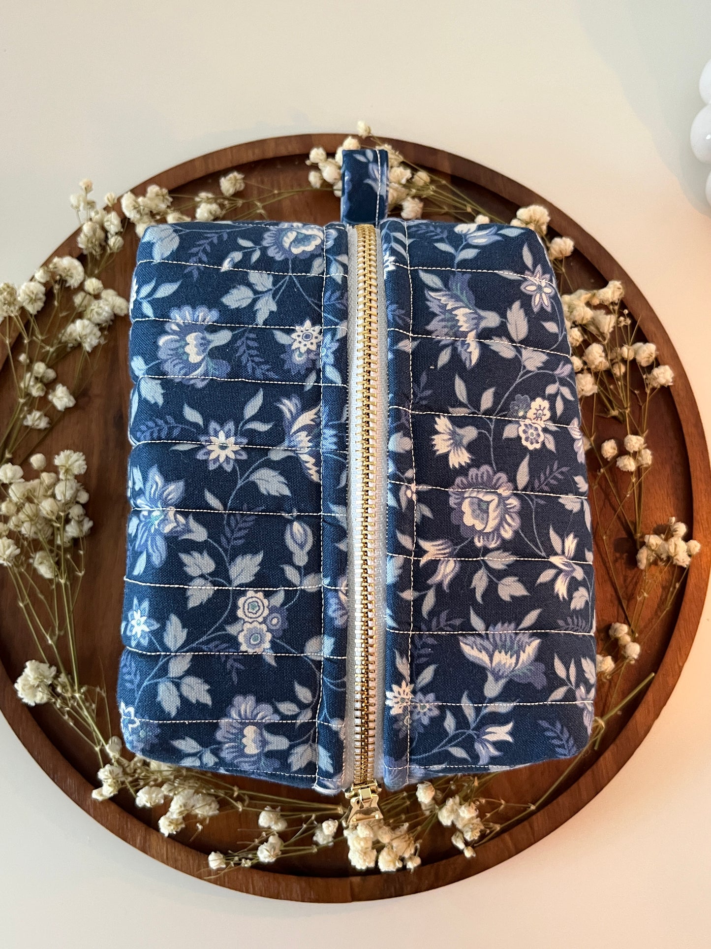 Two blue floral-patterned makeup bags on a wooden tray with dried flowers.