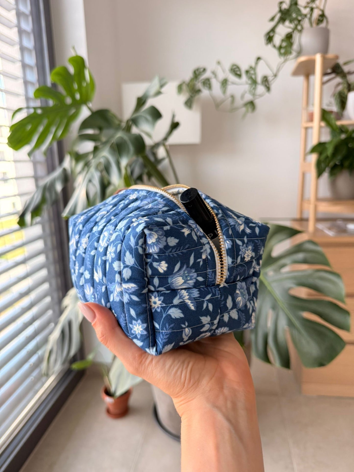 Hand holding a blue floral-patterned makeup bag with plants in the background