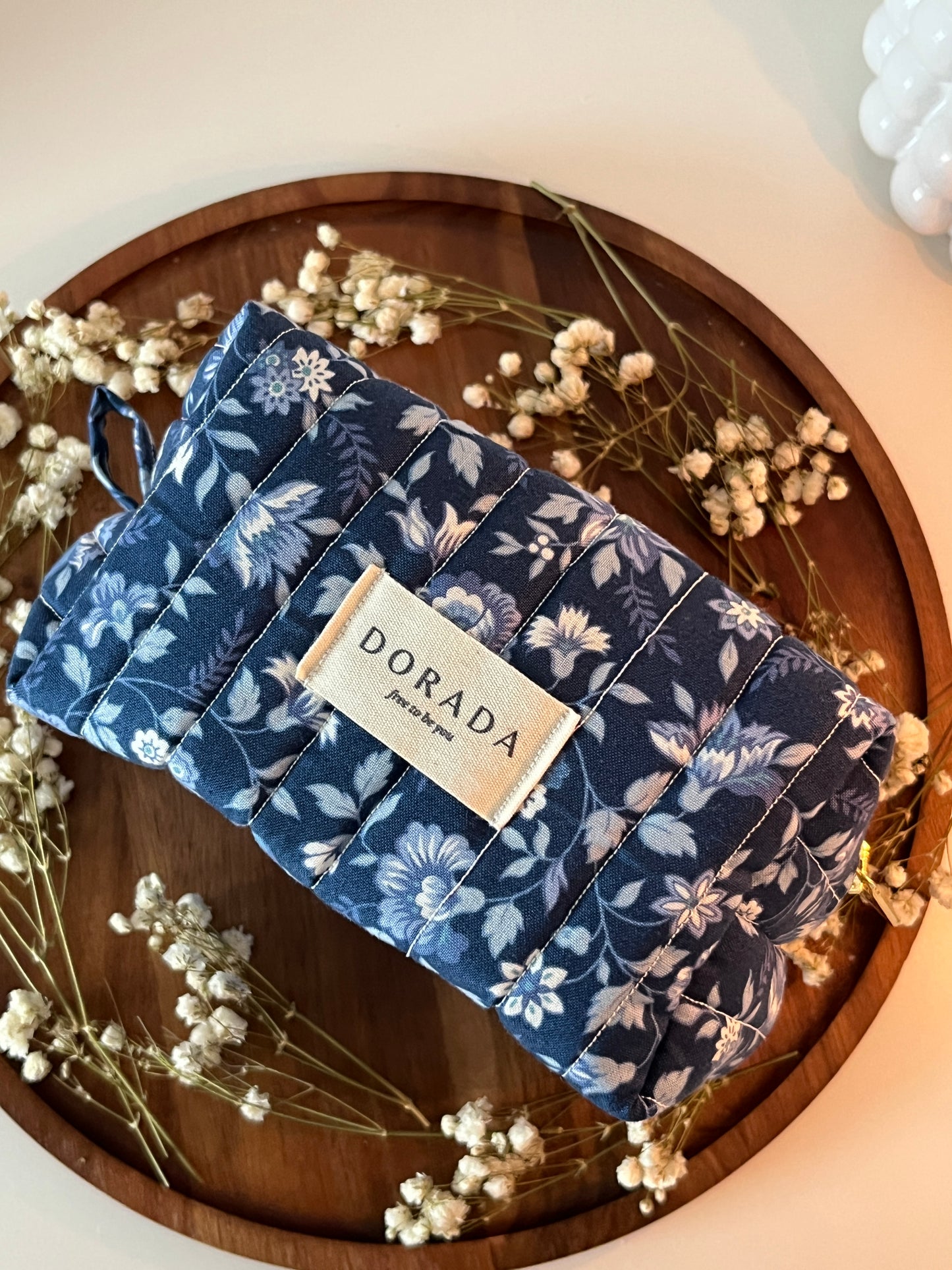 Folded blue floral fabric with a label on a wooden surface surrounded by dried flowers.