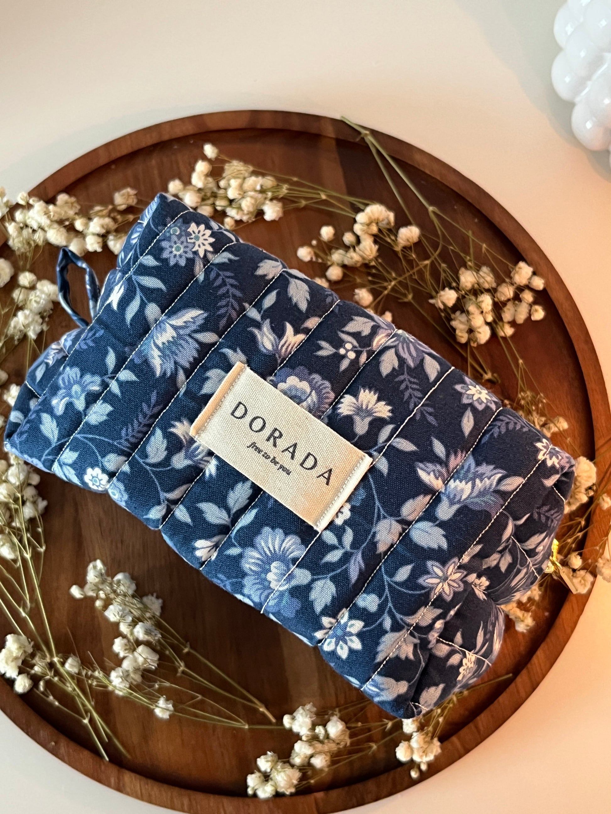 Folded blue floral fabric with a label on a wooden surface surrounded by dried flowers.